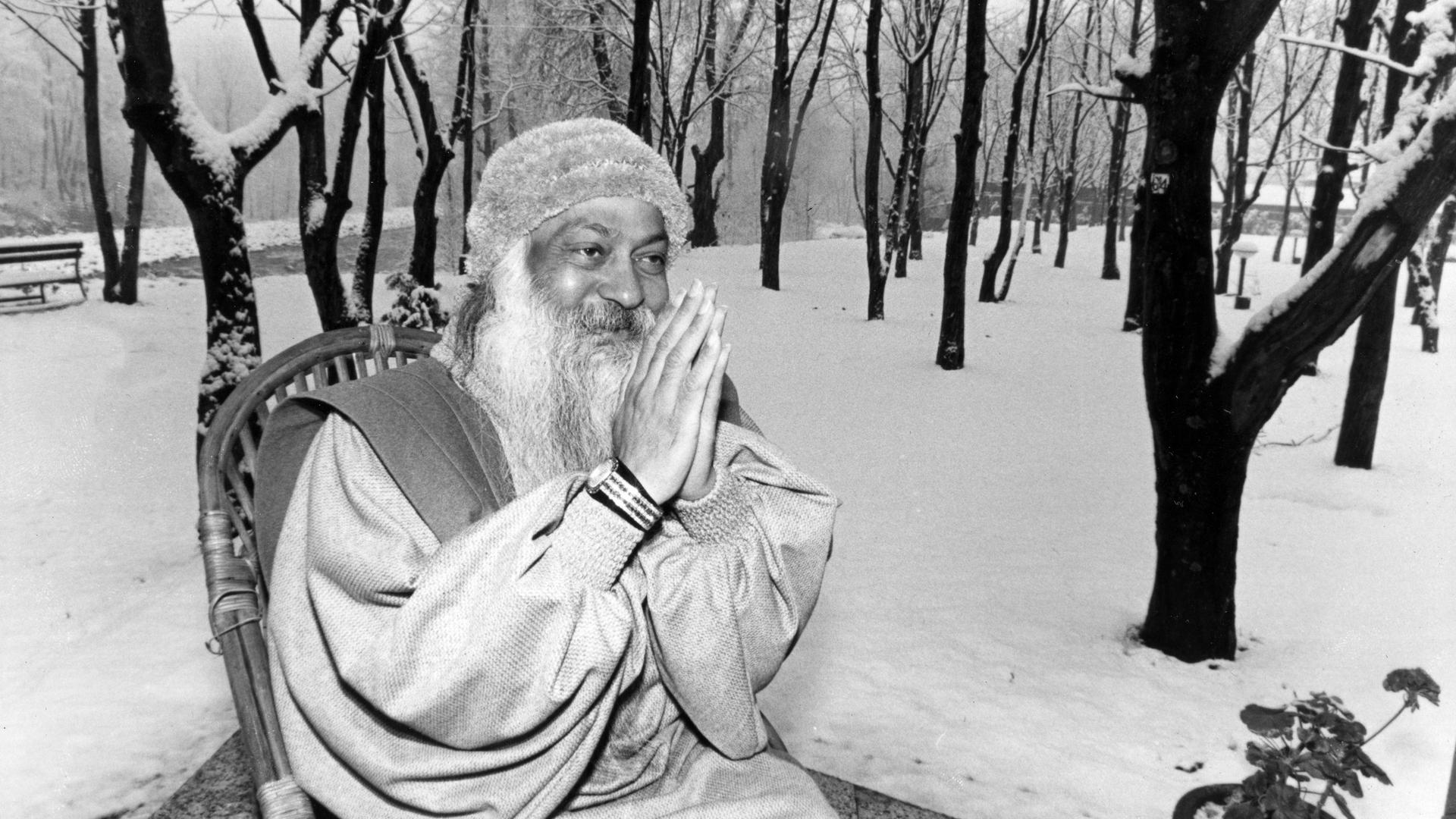 Bhagwan Shree Rajneesh hat die Hände wie zum Gebet zusammengelegt und lächelt. Er sitzt auf einem Stuhl in einer winterlichen Landschaft in Manali (Indien). Die Aufnahme ist vom 27.12.1985.