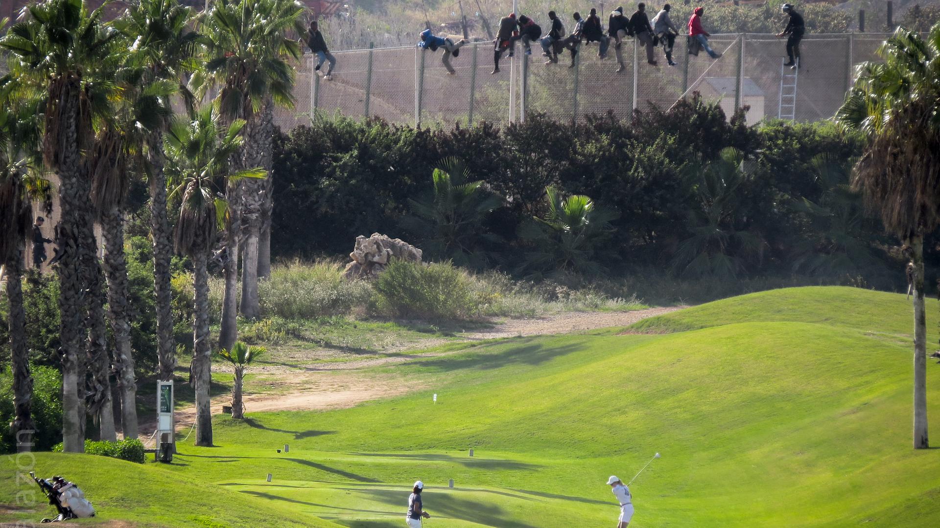 Ein Pärchen spielt unbekümmert Golf, obwohl direkt hinter ihm ein hoher Stacheldrahtzaun aufragt, ein Grenzzaun der spanischen Enklave Melilla in Marokko. Auf dem Zaun erkennt man einige afrikanische Männer, die gerade versuchen, hinüberzuflüchten.