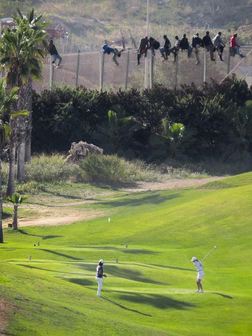 Ein Pärchen spielt unbekümmert Golf, obwohl direkt hinter ihm ein hoher Stacheldrahtzaun aufragt, ein Grenzzaun der spanischen Enklave Melilla in Marokko. Auf dem Zaun erkennt man einige afrikanische Männer, die gerade versuchen, hinüberzuflüchten.