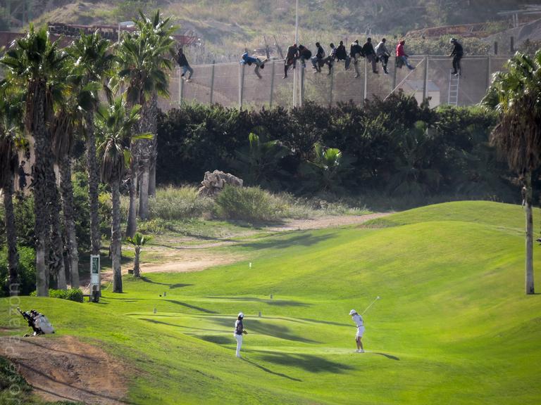 Ein Pärchen spielt unbekümmert Golf, obwohl direkt hinter ihm ein hoher Stacheldrahtzaun aufragt, ein Grenzzaun der spanischen Enklave Melilla in Marokko. Auf dem Zaun erkennt man einige afrikanische Männer, die gerade versuchen, hinüberzuflüchten.