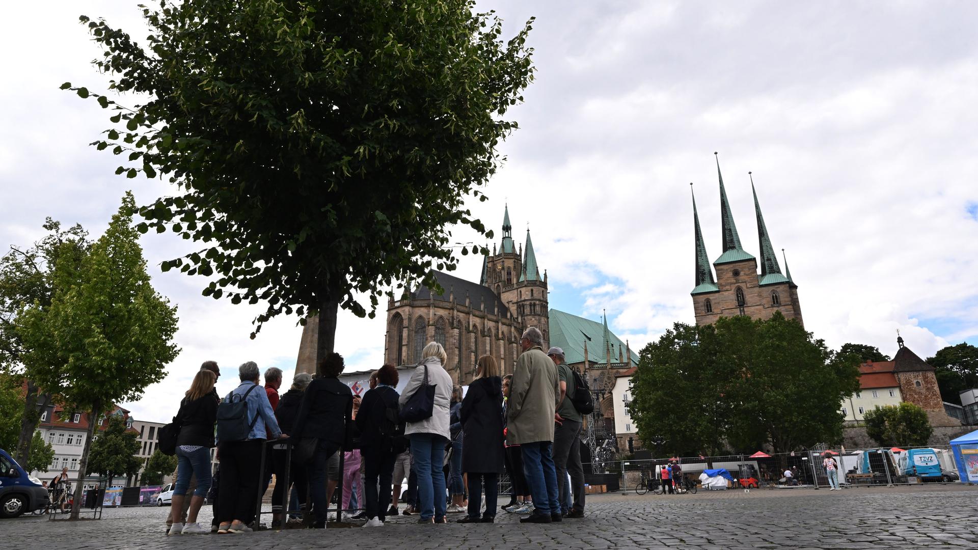 Der Domplatz in Erfurt: Im Vordergrund stehen mehrere Personen vor einem Baum, im Hintergrund sieht man den Dom und eine Kirche. 