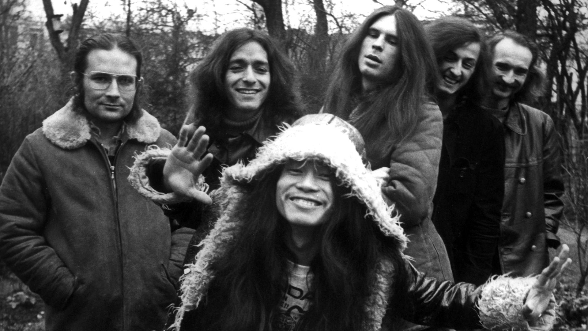 Irmin Schmidt, Jaki Liebezeit, Michael Karoli, Ulli Gerlach, Holger Czukay und  Damo Suzuki von der Band Can in einem Schwarzweißporträt. Die Band entspricht dem typischen Hippie-Bild der frühen Siebziger.