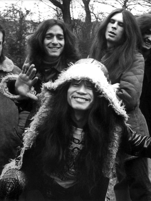 Irmin Schmidt, Jaki Liebezeit, Michael Karoli, Ulli Gerlach, Holger Czukay und  Damo Suzuki von der Band Can in einem Schwarzweißporträt. Die Band entspricht dem typischen Hippie-Bild der frühen Siebziger.