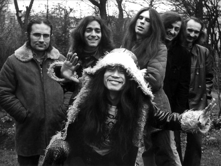 Irmin Schmidt, Jaki Liebezeit, Michael Karoli, Ulli Gerlach, Holger Czukay und  Damo Suzuki von der Band Can in einem Schwarzweißporträt. Die Band entspricht dem typischen Hippie-Bild der frühen Siebziger.