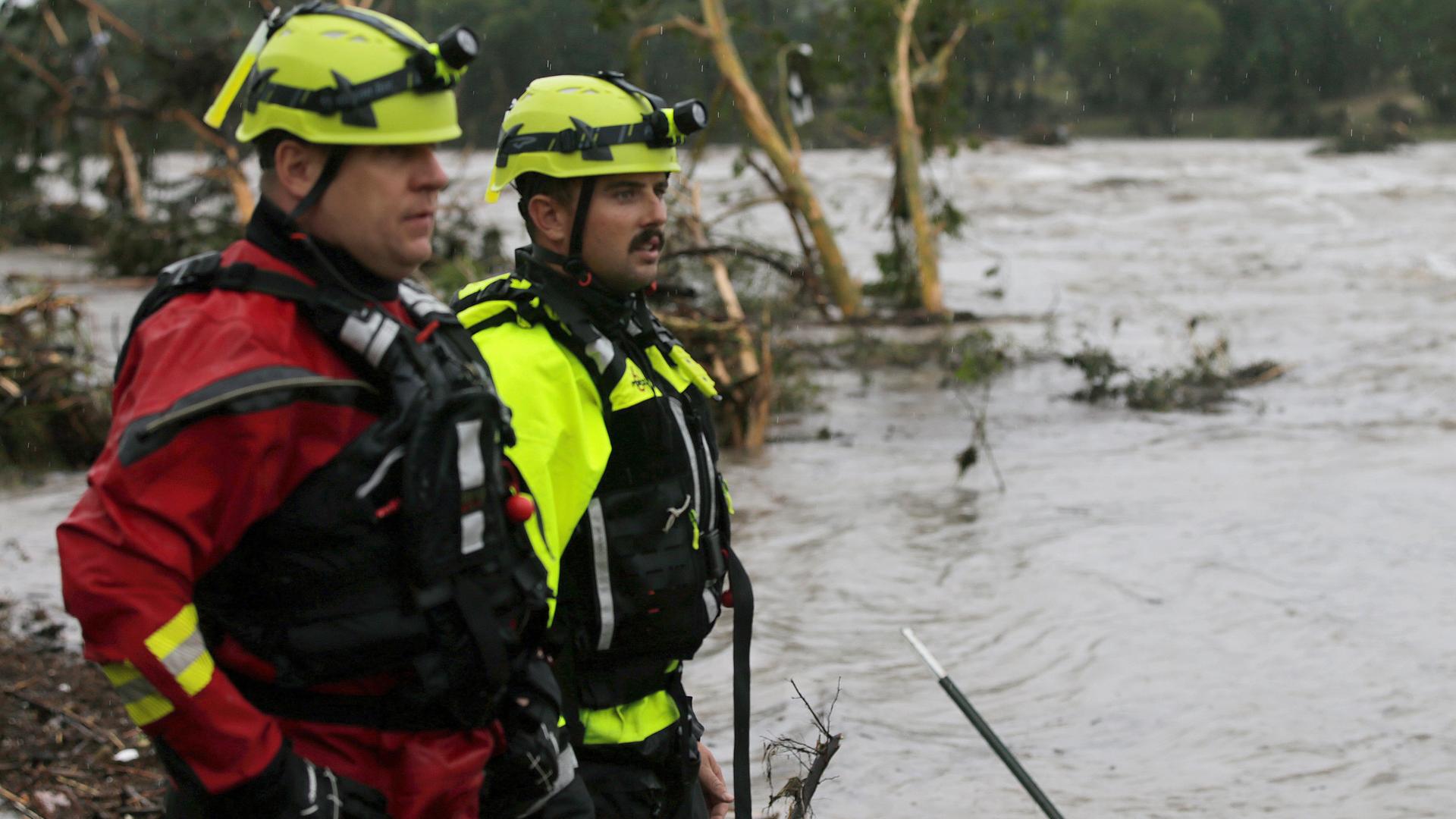 Feuerwehrmänner stehen an einem Flussufer und blicken auf reißende Fluten. Feuerwehrmänner stehen an einem Flussufer und blicken auf reißende Fluten.