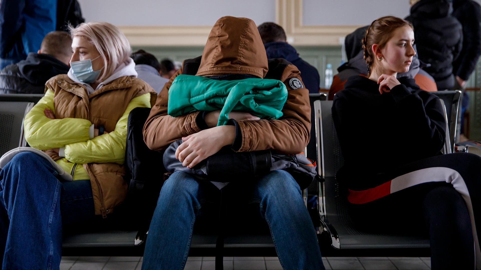 Menschen sitzen in der Wartehalle eines Bahnhofs. Eine Person hat ihren Kopf auf einer Tasche abgelegt.