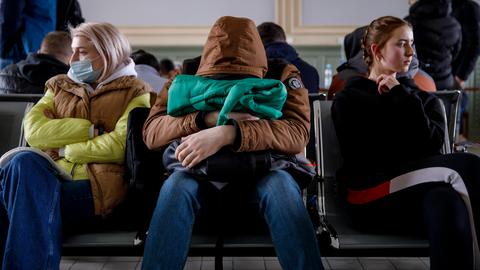 Menschen sitzen in der Wartehalle eines Bahnhofs. Eine Person hat ihren Kopf auf einer Tasche abgelegt.