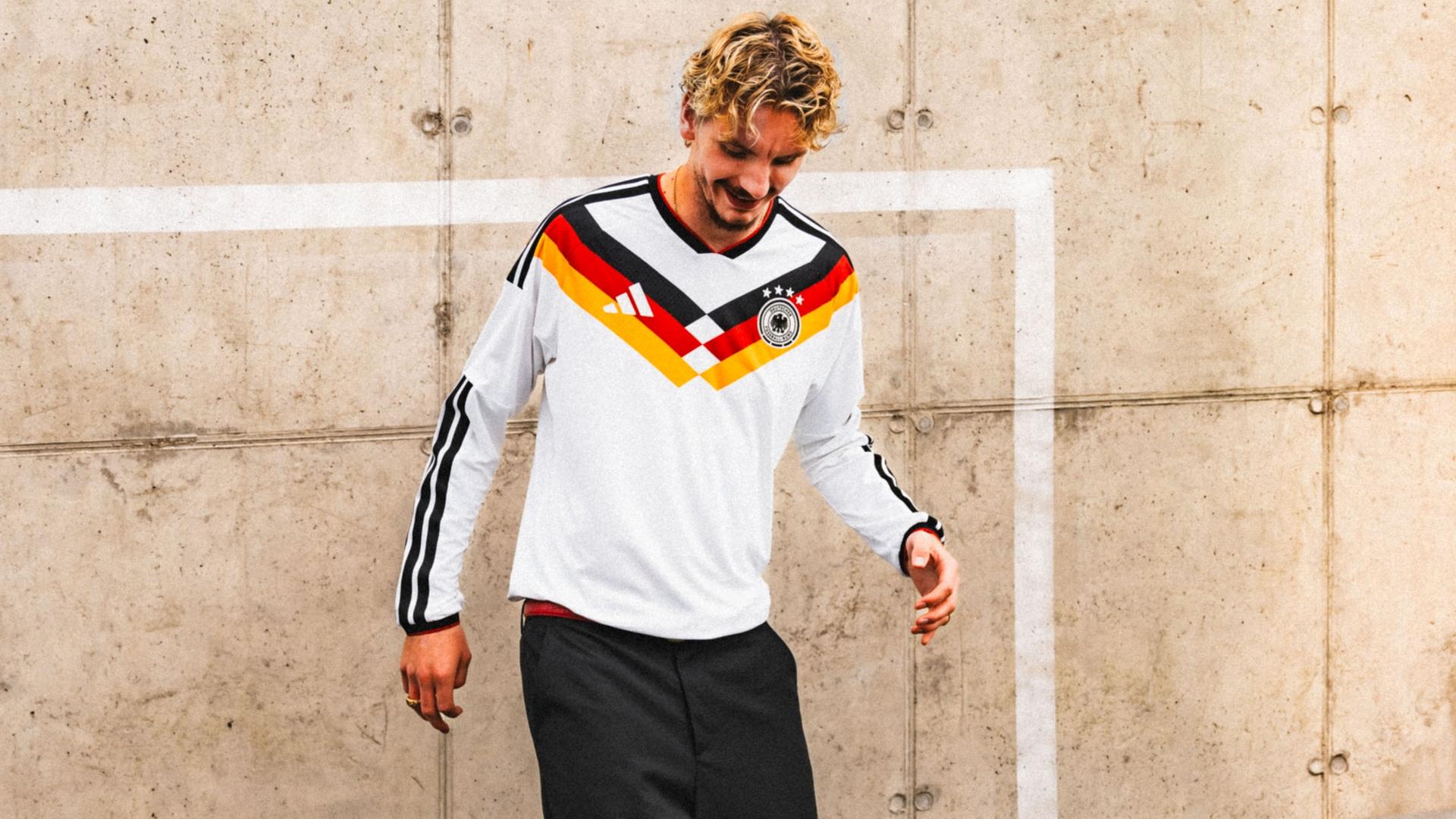 Woltemade mit dem Trikot steht vor einer beigen Wand mit einem aufgemaltem weißen Tor. Er blickt lächelnd nach unten. 