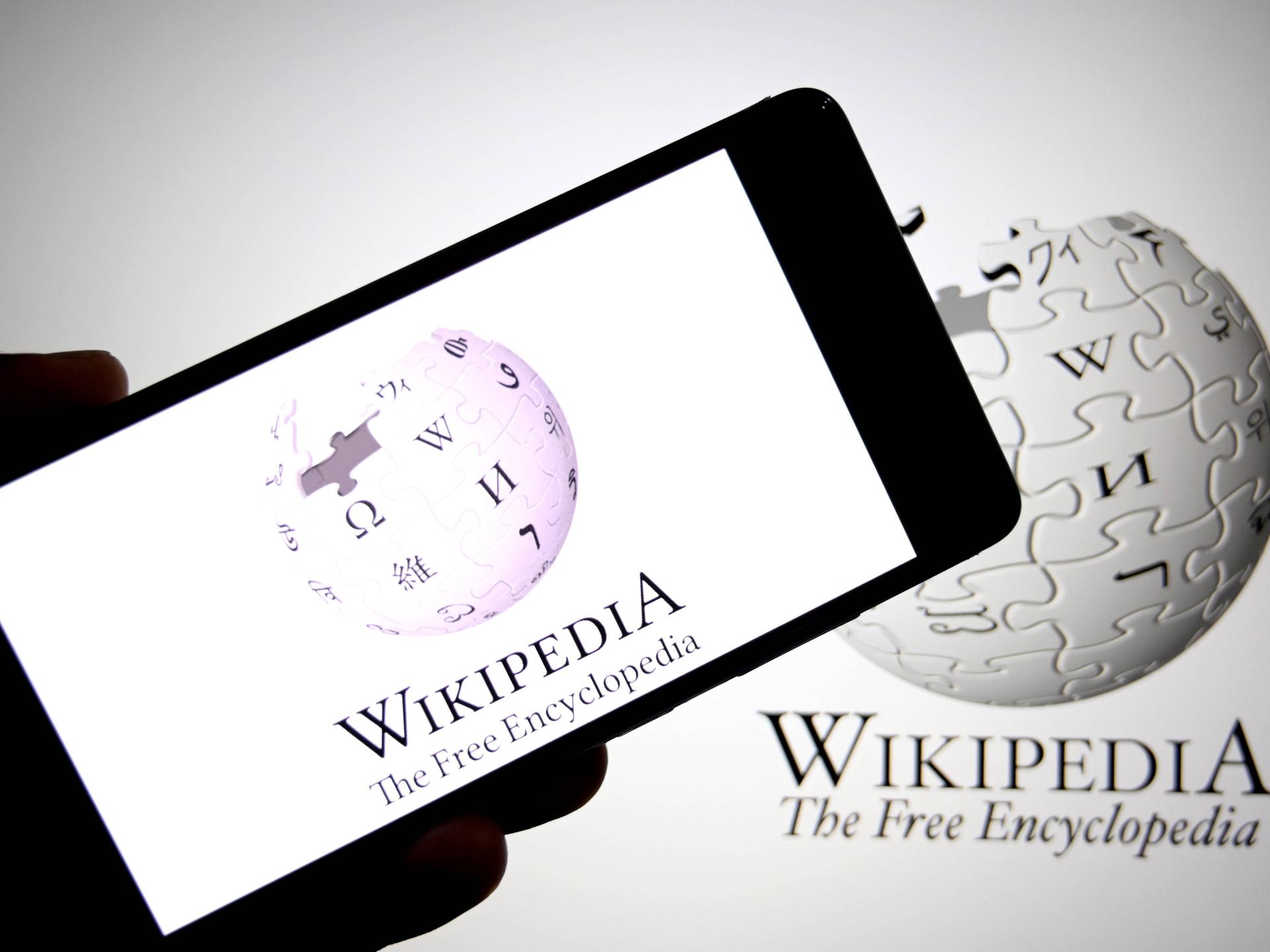 Ein Foto auf dem eine Hand ein Smartphone mit Wikipedia auf dem Display vor einem Bildschirm mit Wikipedia auf dem Displaz hält.