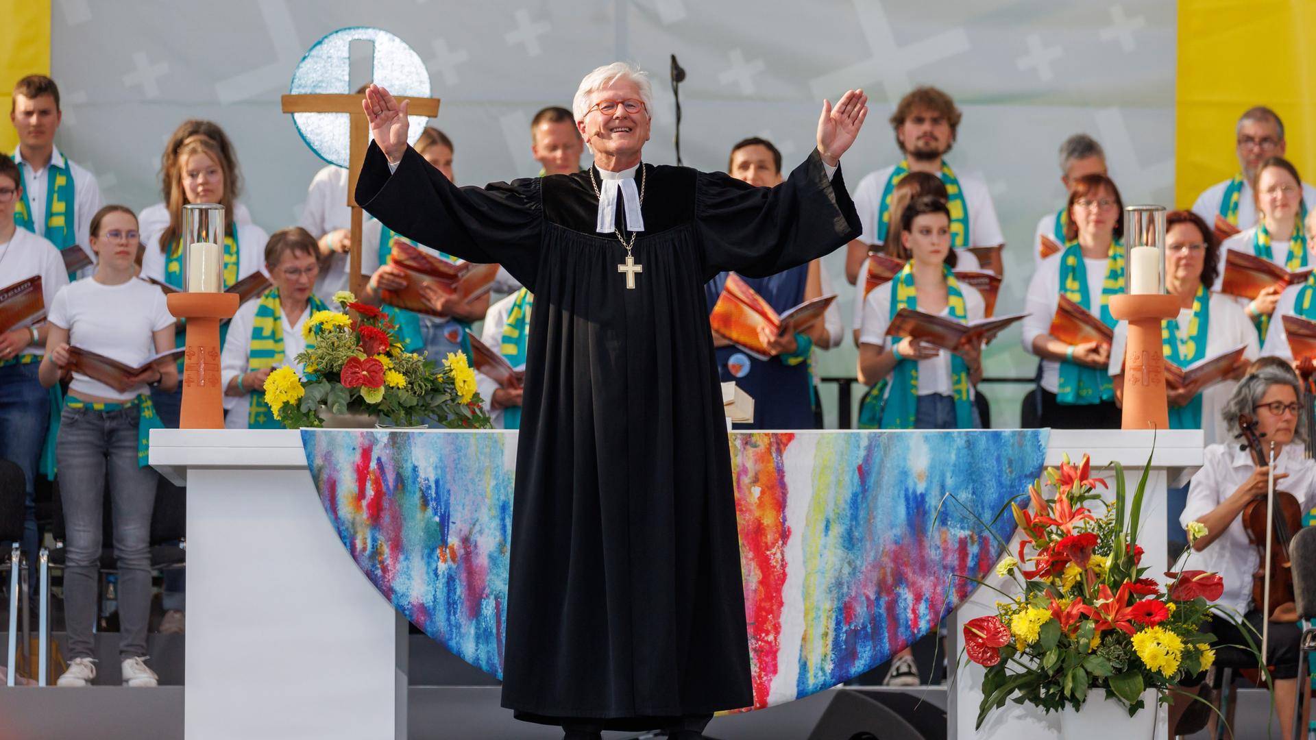 Ein Mann in Kirchen-Kleidung steht auf einer Bühne mit Blumen und anderen Menschen und hebt lachend die Arme zum Segen hoch.