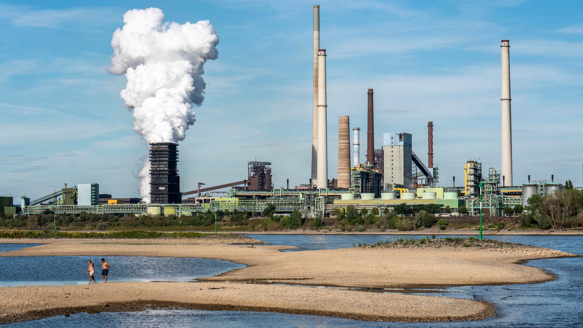 Auf Sandbänken am Rhein gehen zwei Menschen spazieren, im Hintergrund das Stahlwerk Kokerei Schwelgern in Duisburg-Bruckhausen.
Auf Sandbänken am Rhein gehen zwei Menschen spazieren, im Hintergrund das Stahlwerk Kokerei Schwelgern in Duisburg-Bruckhausen.