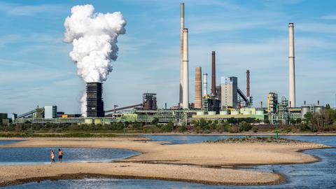 Auf Sandbänken am Rhein gehen zwei Menschen spazieren, im Hintergrund das Stahlwerk Kokerei Schwelgern in Duisburg-Bruckhausen.
      