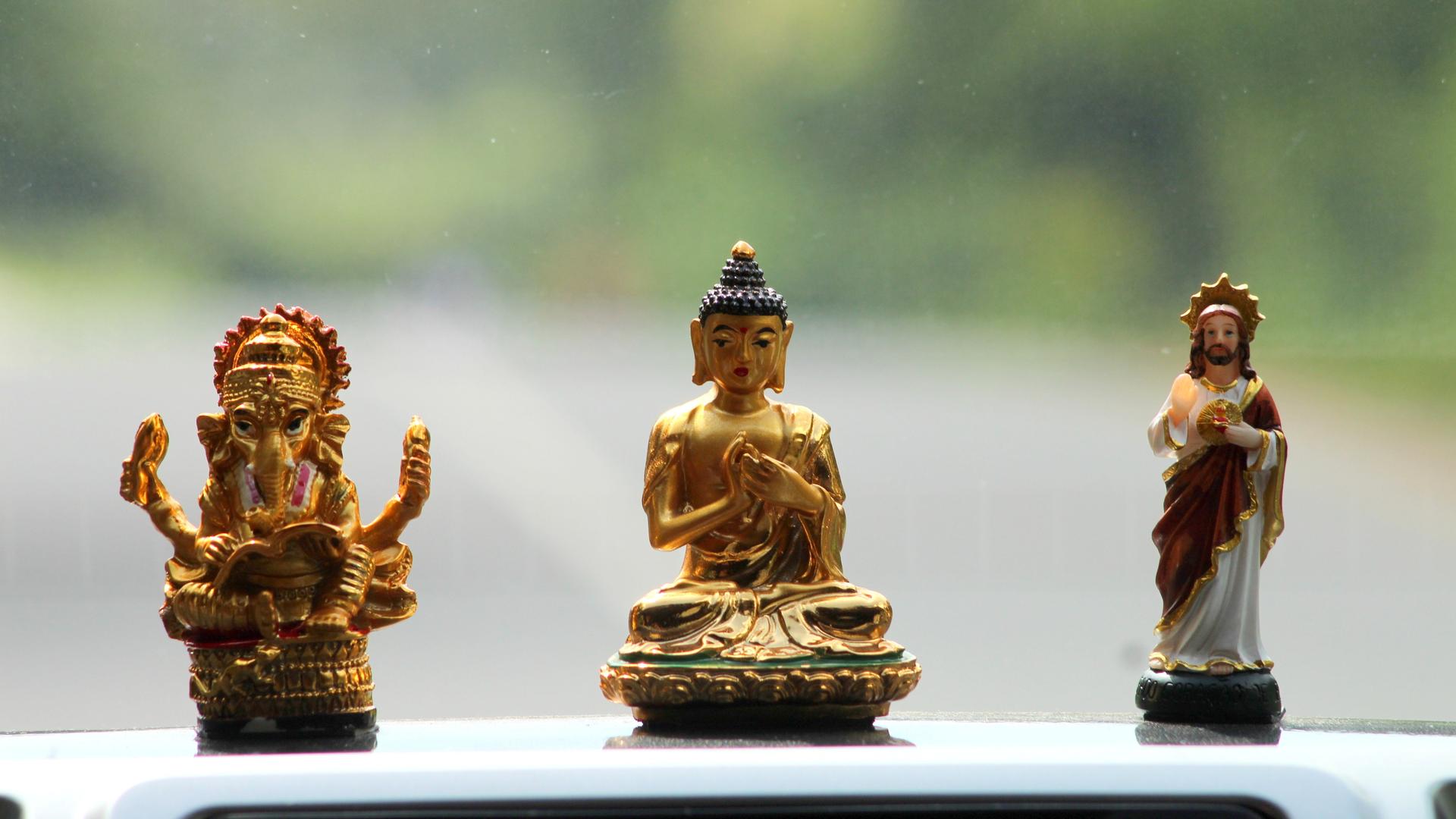Drei Figuren unterschiedlicher Glaubensrichtungen, Hinduismus, Buddhismus und Christentum, stehen auf einem Autodach. Der Hintergrund ist verschwommen grün.