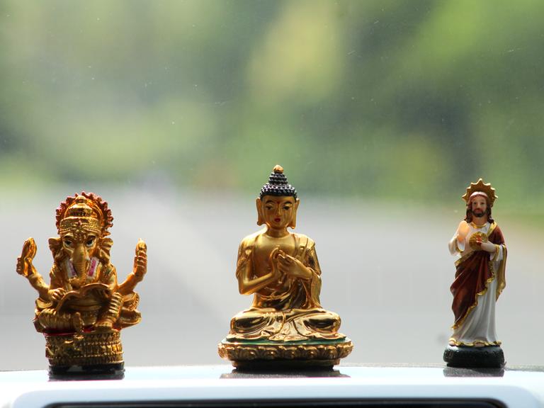Drei Figuren unterschiedlicher Glaubensrichtungen, Hinduismus, Buddhismus und Christentum, stehen auf einem Autodach. Der Hintergrund ist verschwommen grün.