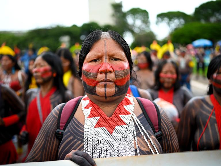Eine indigene Frau steht an der Spitze einer Demonstration. Ihr Gesicht ist in traditionellen Farben bemalt.