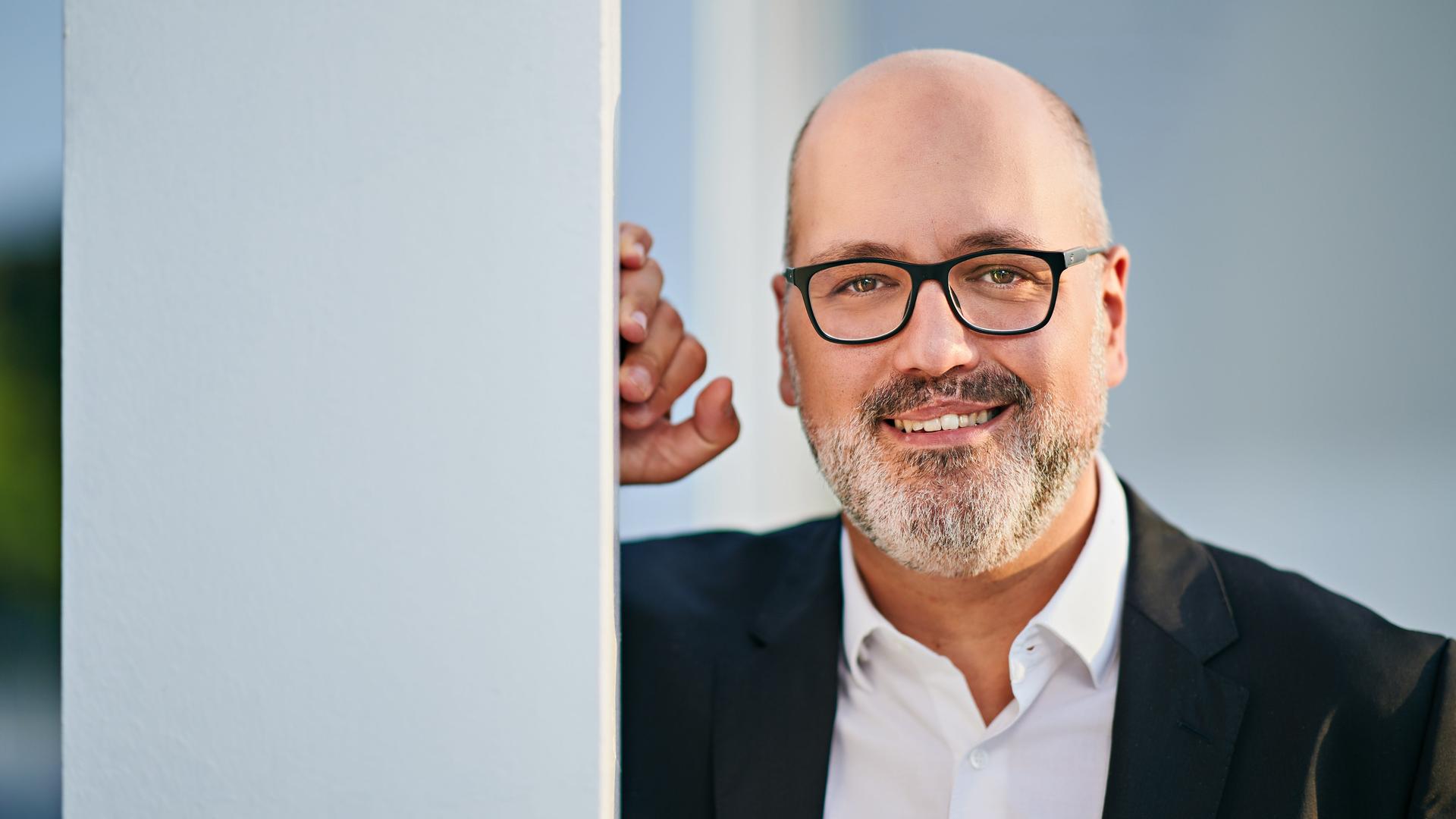 Ein Mann mit Glatze und gräulich meliertem dunklem Bart blickt lächelnd in die Kamera. Mit der rechten Hand stützt er sich an einer weißen Strebe ab. Er trägt ein weißes Hemd und ein schwarzes Jackett.