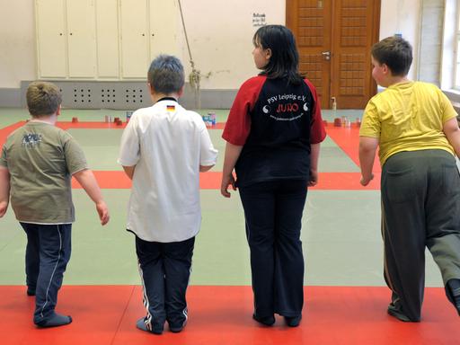 Vier Kinder mit Übergewicht stehen in einer Turnhalle auf einer Matte mit dem Rücken zum Betrachter. Ein Junge rechts hebt das rechte Bein nach hinten. Vier Kinder mit Übergewicht stehen in einer Turnhalle auf einer Matte mit dem Rücken zum Betrachter. Ein Junge rechts hebt das rechte Bein nach hinten.