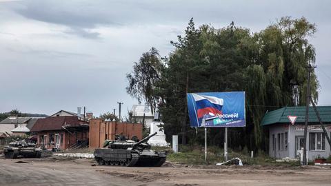 Ein zurück gelassener russischer Panzer auf einer zerstörten Straße in Izjum, Ukraine. Im Hintergrund  ist ein Plakat mit der Flagge Russlands zu sehen.