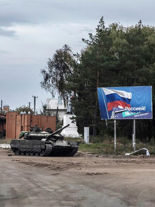 Ein zurück gelassener russischer Panzer auf einer zerstörten Straße in Izjum, Ukraine. Im Hintergrund  ist ein Plakat mit der Flagge Russlands zu sehen.