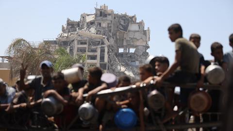 Palästinenser, darunter auch Kinder versammeln sich in Gaza-Stadt, um warme Mahlzeiten entgegenzunehmen, die von der Wohltätigkeitsorganisation verteilt werden. Im Hintergrund ist ein zerstörtes Gebäude zu sehen.