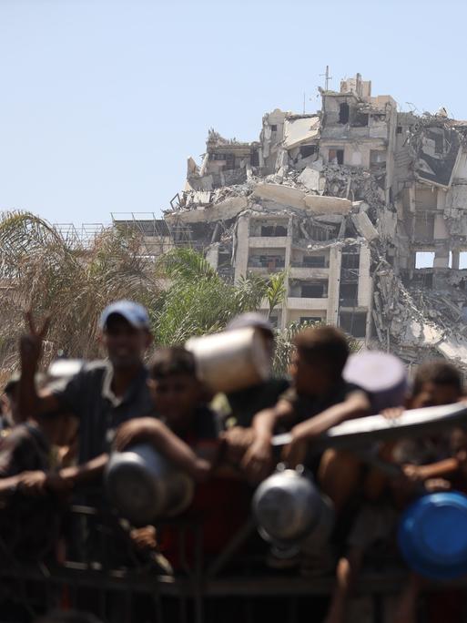 Palästinenser, darunter auch Kinder versammeln sich in Gaza-Stadt, um warme Mahlzeiten entgegenzunehmen, die von der Wohltätigkeitsorganisation verteilt werden. Im Hintergrund ist ein zerstörtes Gebäude zu sehen.