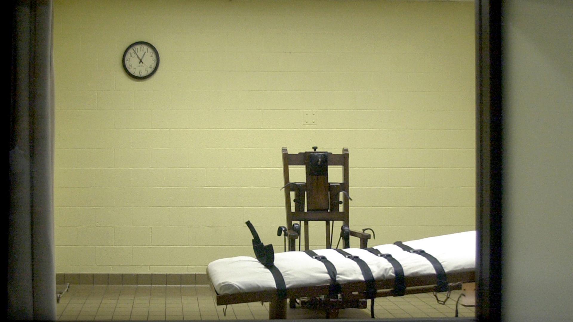 Blick vom Zeugenraum aus in die Hinrichtungskammer einer amerikanischen Justizvollzugsanstalt, in der sich ein Stuhl und eine Liege mit Gurten befindet.