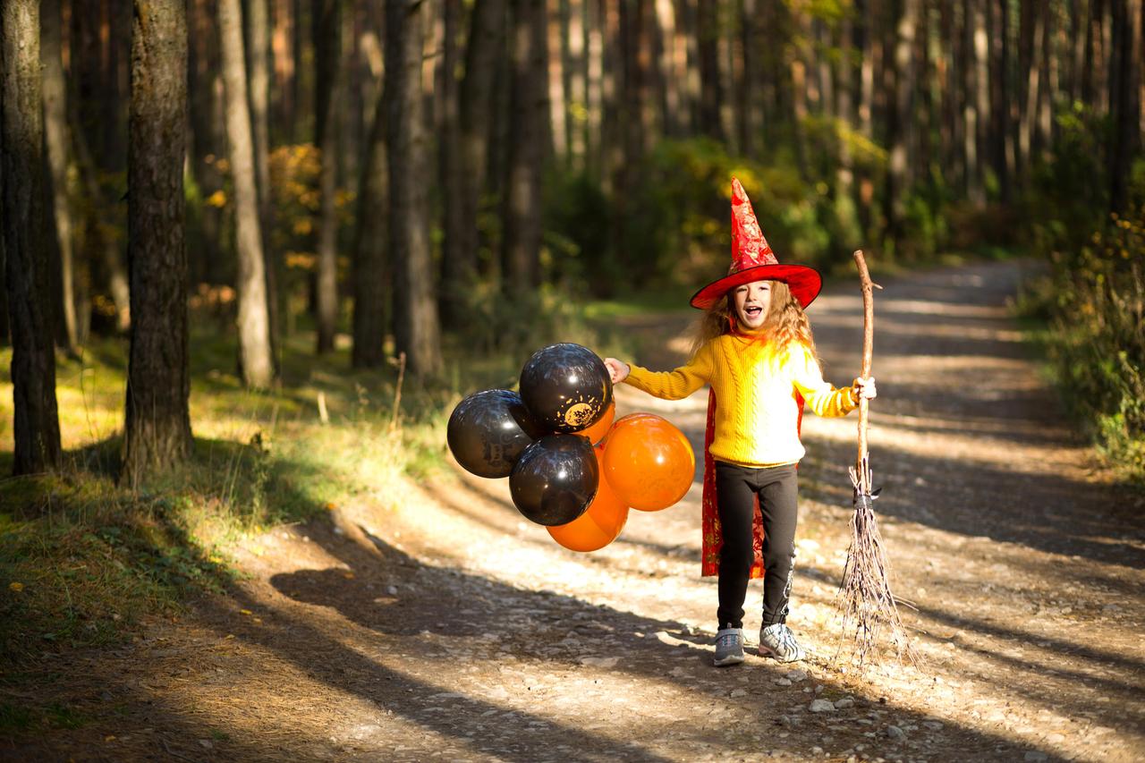 Ein Mädchen in einem Hexenkostüm und mit Luftballons in der Hand im Herbstwald unterwegs zu einer Halloween Party.