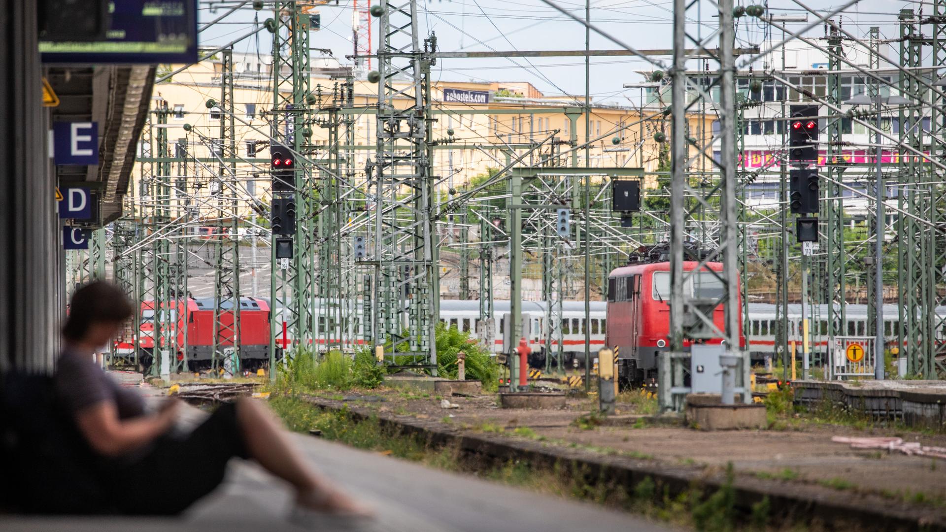 Eine Frau sitzt an einem Bahnsteig des Stuttgarter Hautpbahnhofs. Im Hintergrund sind Züge und Oberleitungen zu sehen. 