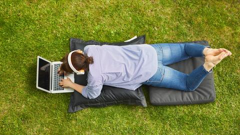 Frau am Laptop Computer bei Videokonferenz liegend auf einer Wiese. (Symbolbild)