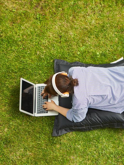 Frau am Laptop Computer bei Videokonferenz liegend auf einer Wiese. (Symbolbild)