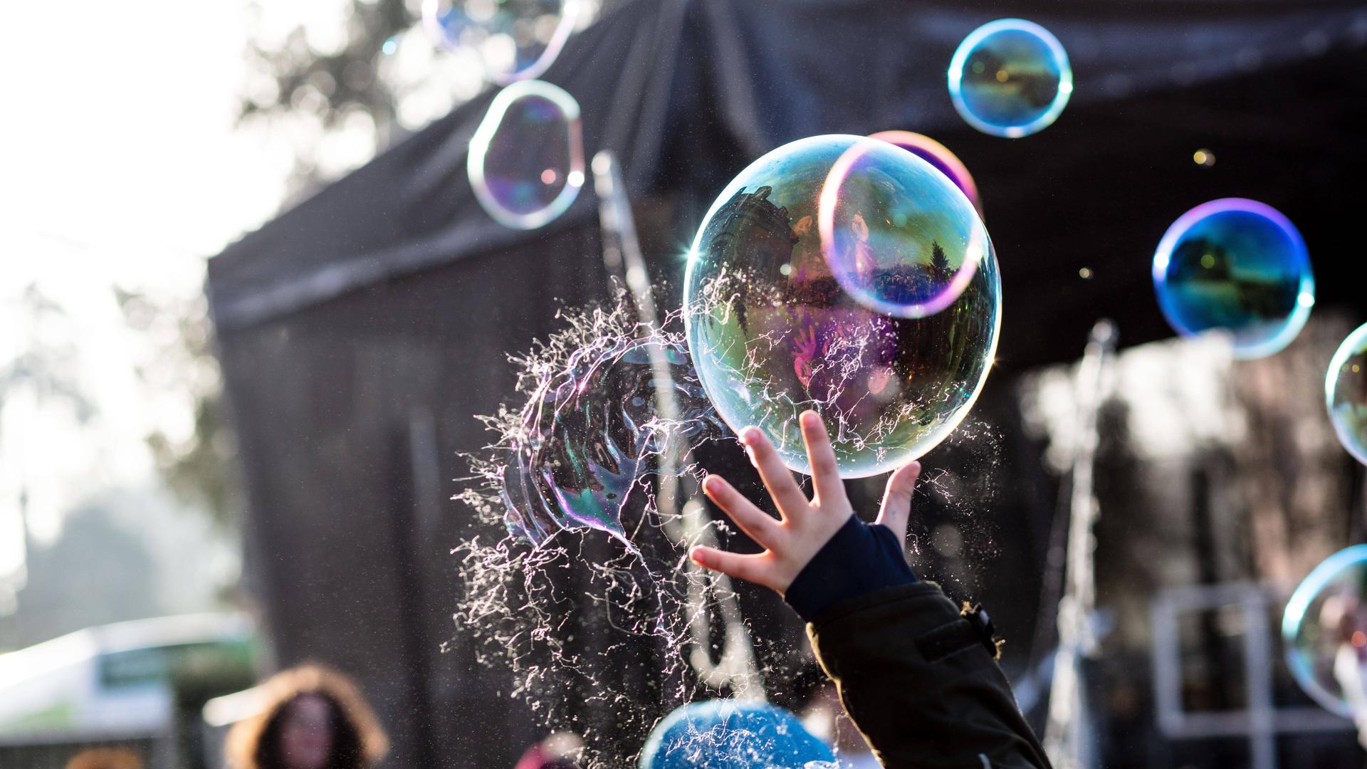 Viele Seifenblasen fliegen in der Luft. Eine Hand lässt eine Blase platzen.