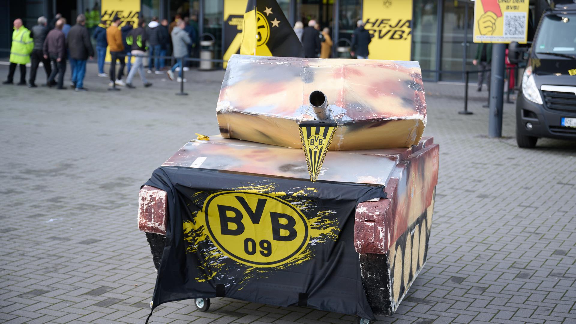 Der Papp-Panzer auf Rädern steht vor der Halle. Am Abschussrohr hängt ein BVB-Wimpel, auf dem Panzer klebt das schwarz-gelbe Vereinsemblem. Im Hintergrund betreten Teilnehmer die Halle.