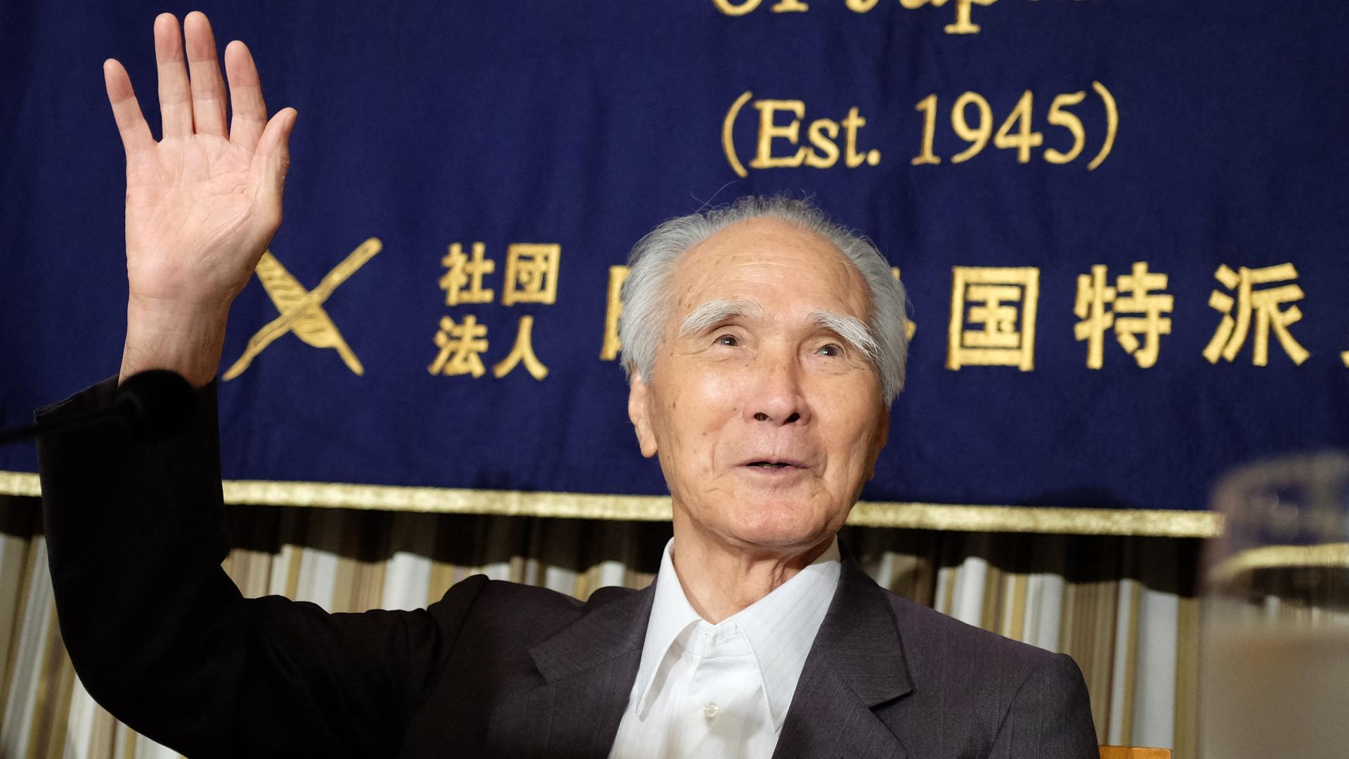Murayama winkt, im Hintergrund eine blaue Fahne mit chinesischen Zeichen und man kann noch "(Est. 1945)" lesen.