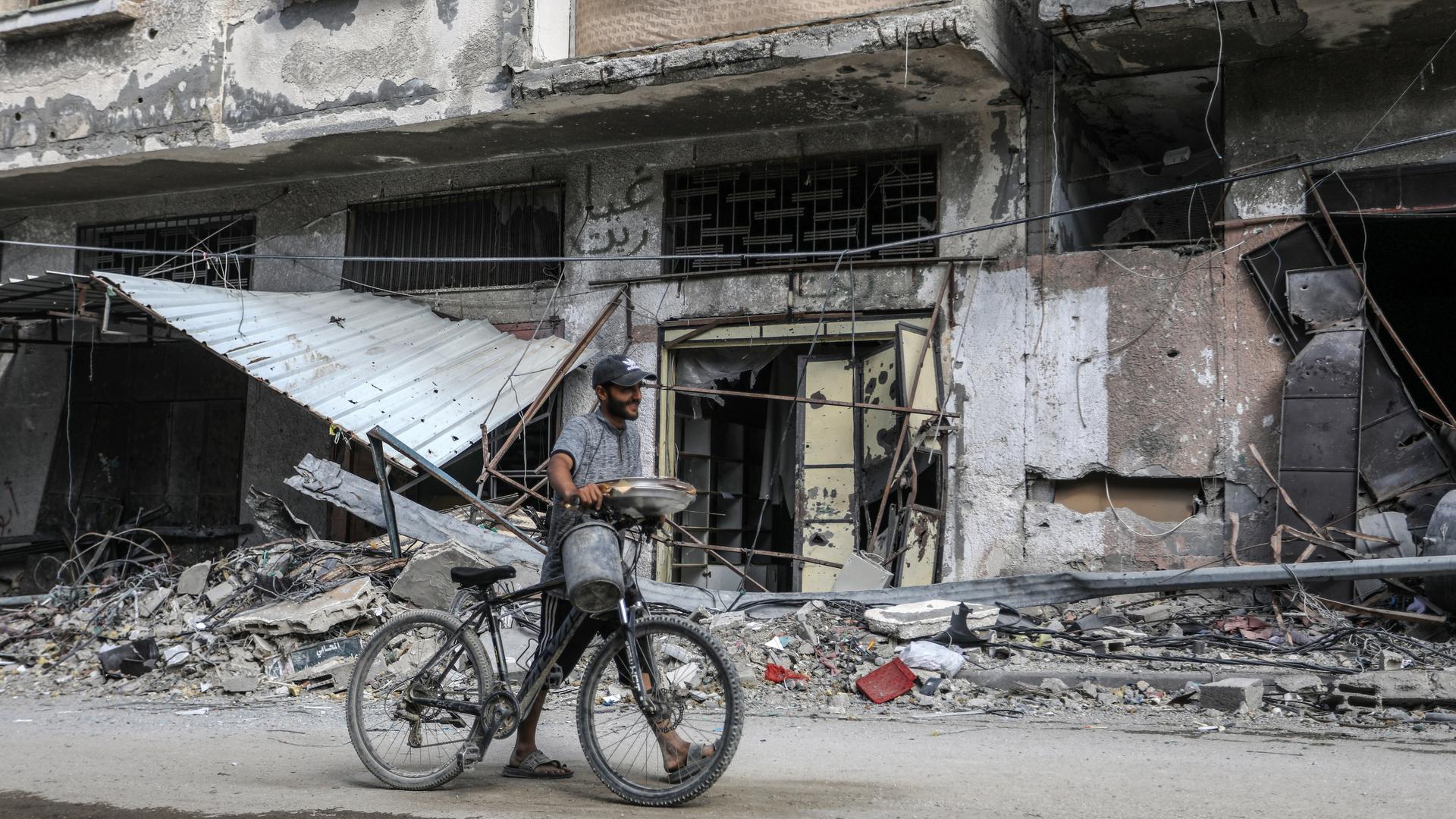 Eine zerstörte Häuserfront in Chan Junis, davor ein Mann mit Fahrrad und Broten in einem silbernen Behältnis in der Hand. 