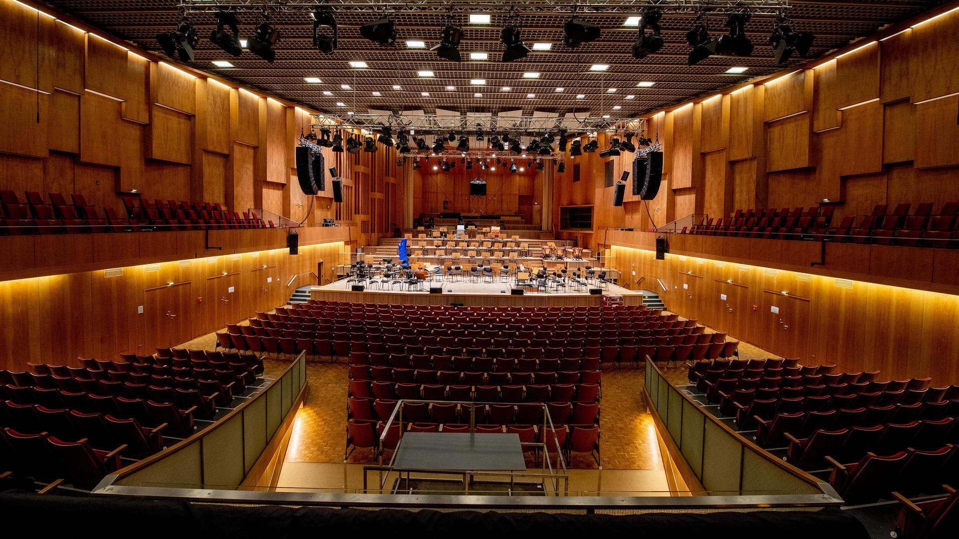 Ein Konzertsaal mit holzvertäfelten Wänden, in dem rote Stühle zu sehen sind sowie eine Bühne, die für ein Orchester ausgestattet ist. Von der Decke hängen Scheinwerfer und Lautsprecherboxen.