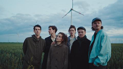 Die Band steht auf einem grünen Feld in Herbskleidung, im Hintergrund erscheinen Windräder.