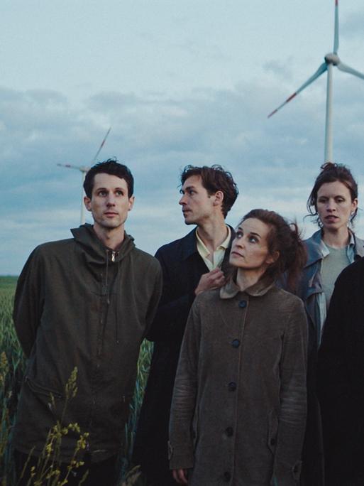 Die Band steht auf einem grünen Feld in Herbskleidung, im Hintergrund erscheinen Windräder.