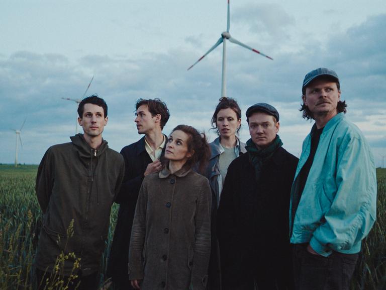 Die Band steht auf einem grünen Feld in Herbskleidung, im Hintergrund erscheinen Windräder.