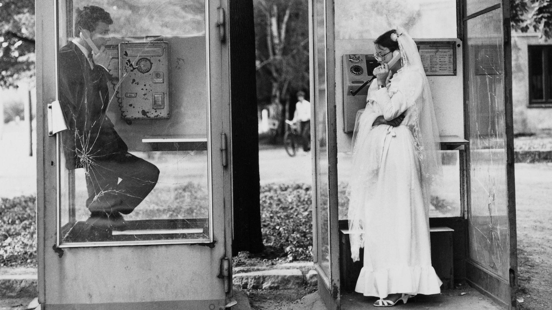 Ein Hochzeitspaar in zwei Telefonzellen in Leipzig, sie telefonieren nebeneinander miteinander, DDR, 1984.