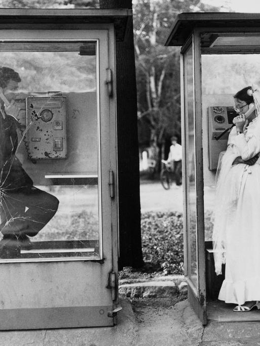 Ein Hochzeitspaar in zwei Telefonzellen in Leipzig, sie telefonieren nebeneinander miteinander, DDR, 1984.