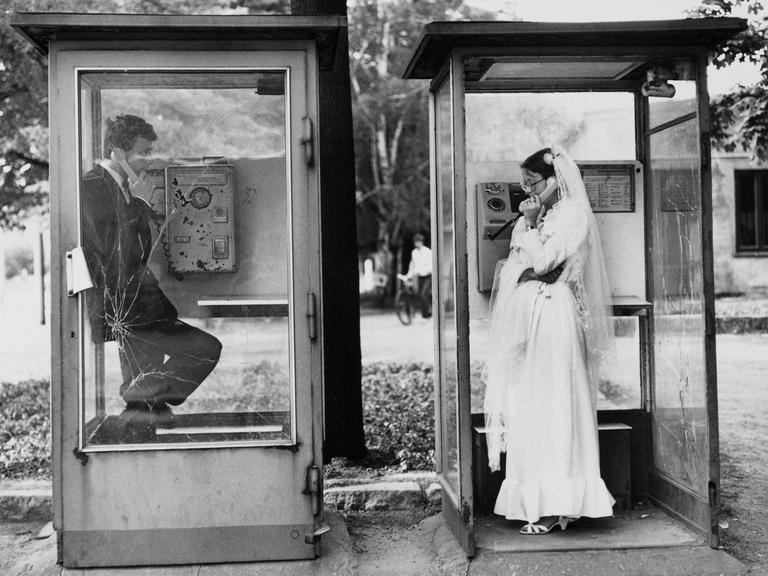 Ein Hochzeitspaar in zwei Telefonzellen in Leipzig, sie telefonieren nebeneinander miteinander, DDR, 1984.