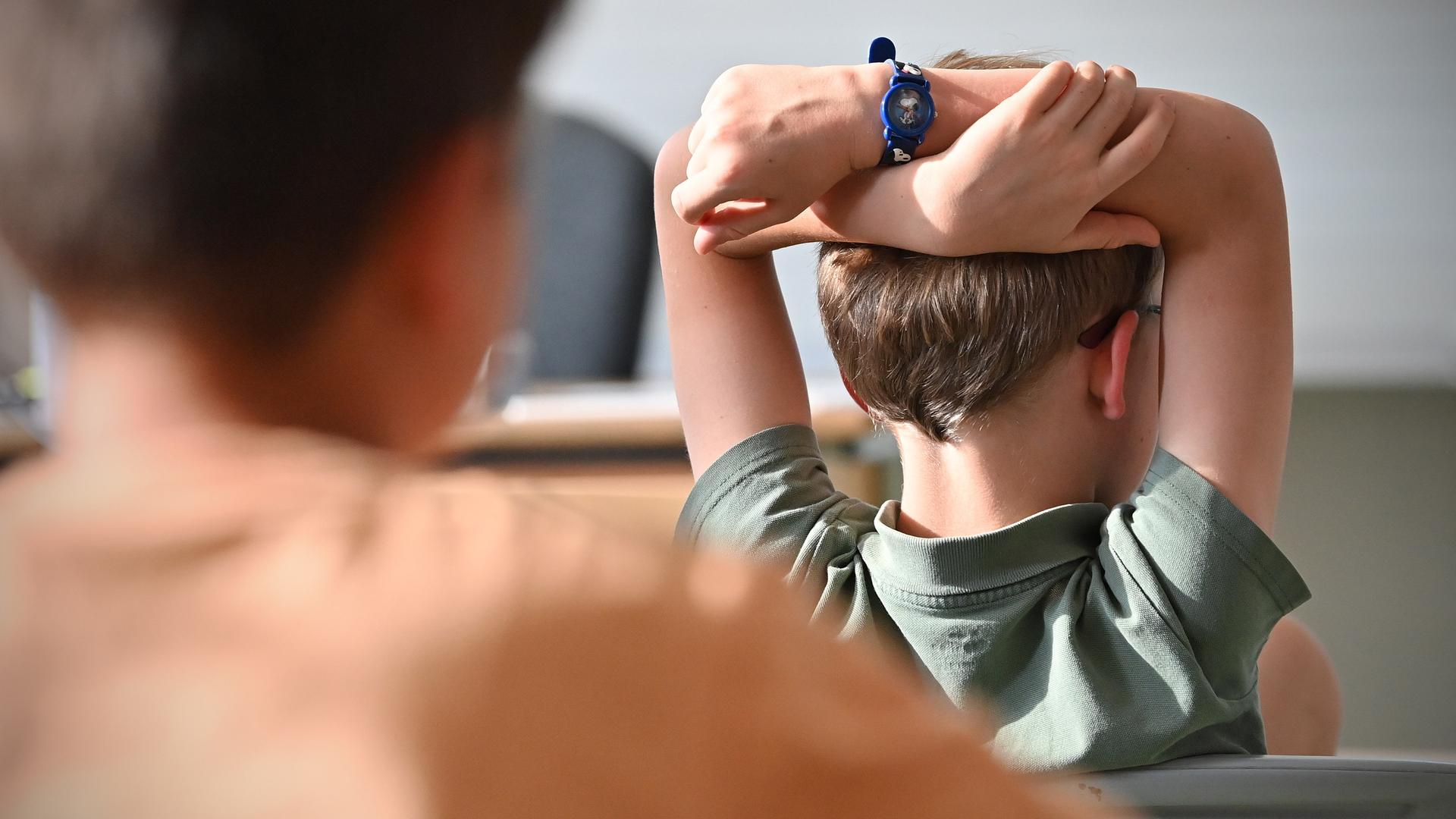 Rückseitiger Blick auf ein Kind was in einem Klassenzimmer sitzt und nach vorn schaut. Es hält die Arme über dem Kopf verschränkt.