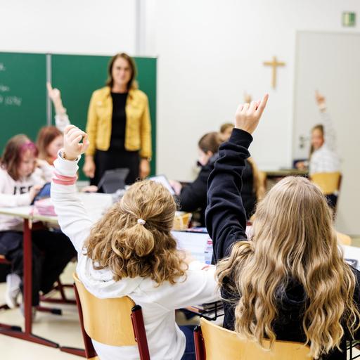 Zwei Schülerinnen zeigen in einer Klasse auf, dahinter steht eine Lehrerin vor der Tafel.