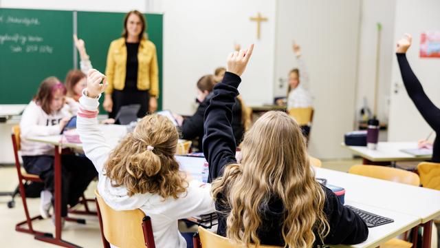 Zwei Schülerinnen zeigen in einer Klasse auf, dahinter steht eine Lehrerin vor der Tafel.