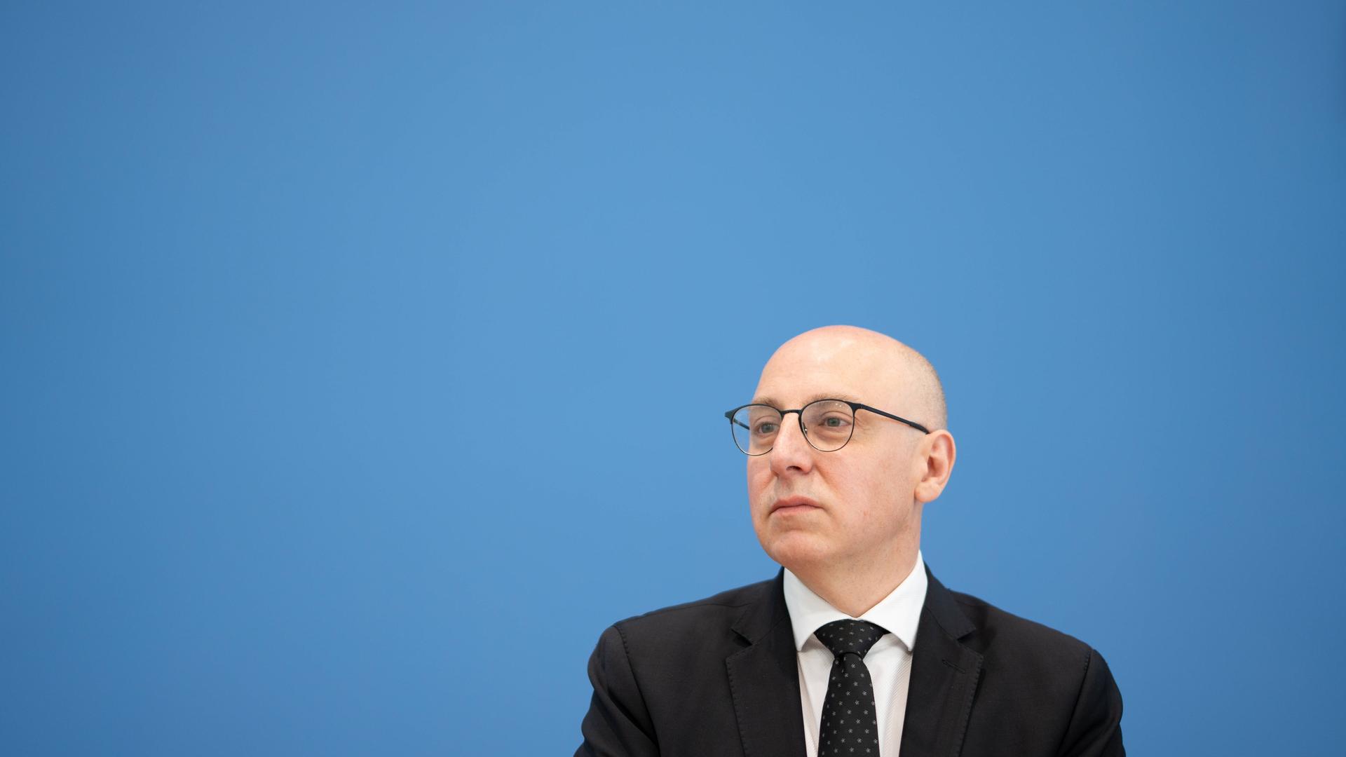 Sinan Selen sitzt im Anzug und mit Brille vor der blauen Wand der Bundespressekonferenz und schaut zum linken Bildrand. Sinan Selen sitzt im Anzug und mit Brille vor der blauen Wand der Bundespressekonferenz und schaut zum linken Bildrand.