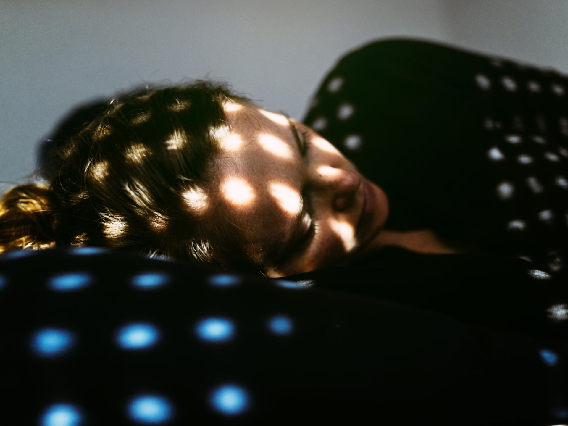 Eine Frau liegt im Dunkeln auf einem Kopfkissen, in ihrem Gesicht sind Lichtpunkte zu sehen, die offensichtlich durch eine Jalousie scheinen