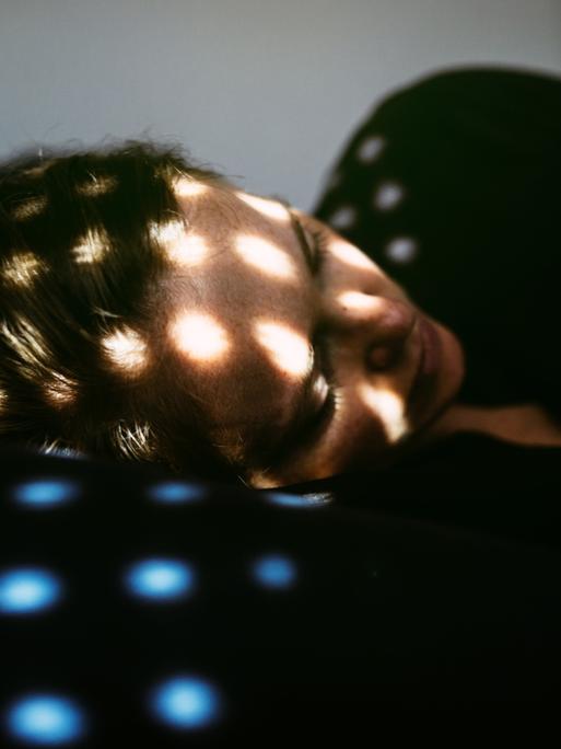 Eine Frau liegt im Dunkeln auf einem Kopfkissen, in ihrem Gesicht sind Lichtpunkte zu sehen, die offensichtlich durch eine Jalousie scheinen
