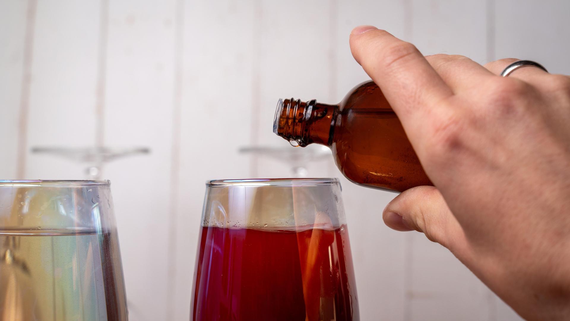 Eine Hand gießt aus einer kleinen braunen Flasche etwas in ein Glas mit einem Getränk.