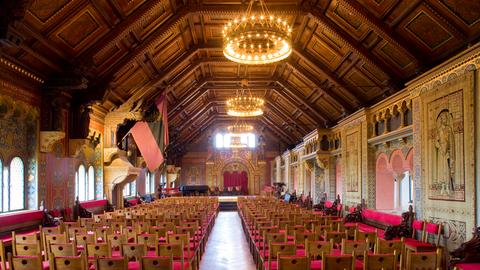 Blick in einen großen, historischen Saal mit hoher, dekorierter Holzdecke, imposanten Leuchten und roten Stühlen.