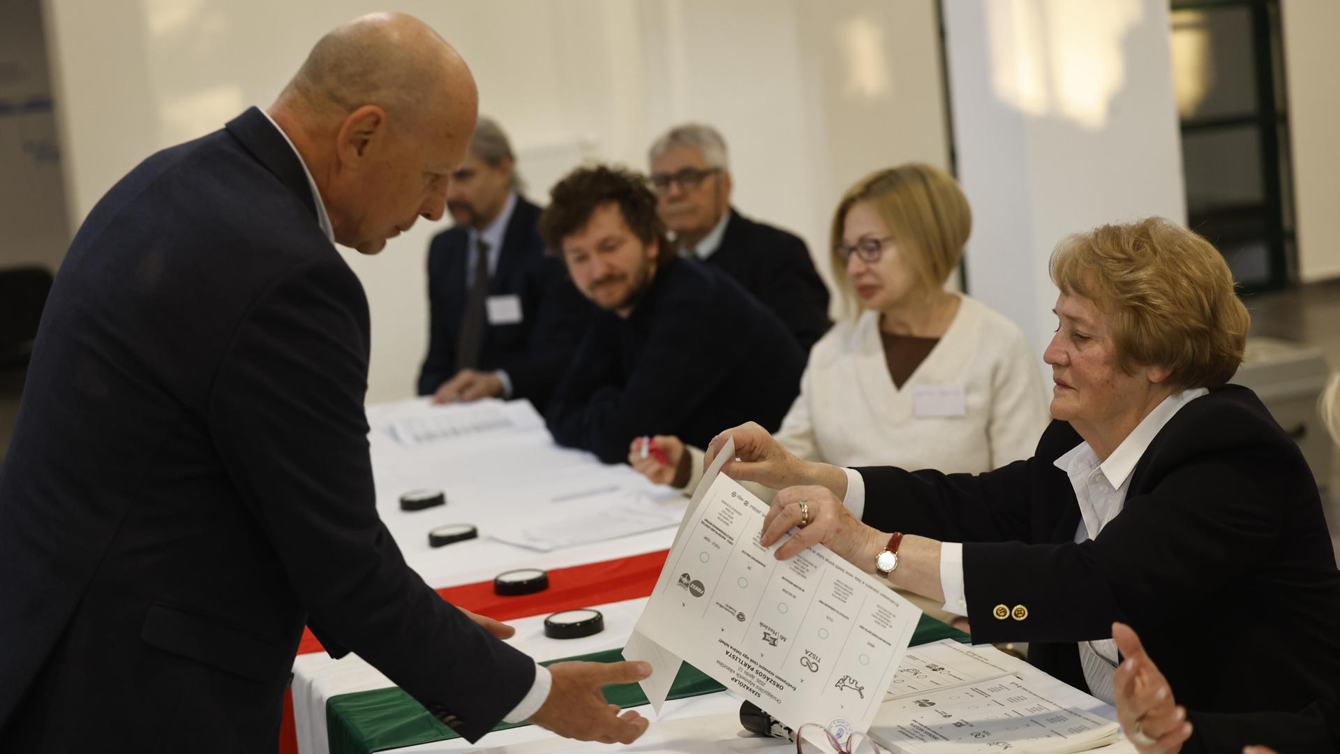 Ein Mann steht vor einem langen Tisch, an dem mehrere Menschen sitzen. Eine Wahlhelferin reicht ihm einen Wahlzettel. Ein Mann steht vor einem langen Tisch, an dem mehrere Menschen sitzen. Eine Wahlhelferin reicht ihm einen Wahlzettel.